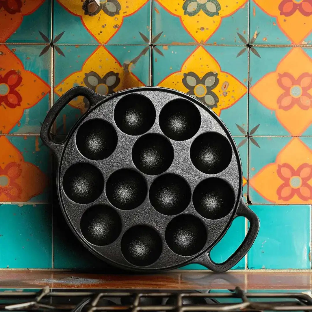 Black cast iron 12-pit Paniyarakkal pan with two handles, leaning against a colorful tiled wall.