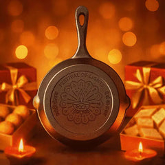 A decorative cast-iron skillet with a handle, featuring an embossed lotus design and the text 'FESTIVAL OF LIGHTS' and 'SACA' on the bottom. The skillet is photographed in a warm, festive setting with glowing candles, gift boxes, and bokeh lights in the background.