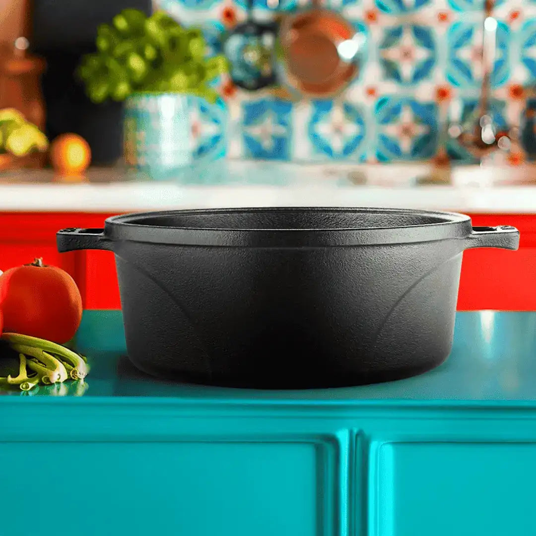 A black cast iron Dutch oven with side handles sits on a bright turquoise countertop in a kitchen setting, with a red tomato, green beans, and a patterned tile backsplash blurred in the background.